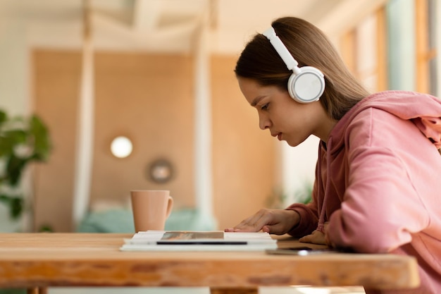 Chica adolescente enfocada inteligente escuchando música en auriculares inalámbricos y leyendo un libro sentado en el escritorio en casa