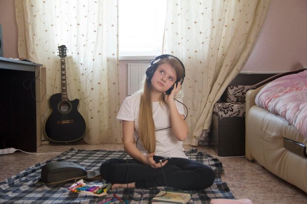 Foto chica adolescente escuchando música en auriculares con teléfono inteligente y dibujo sentado en el suelo en casa, pasatiempo juvenil