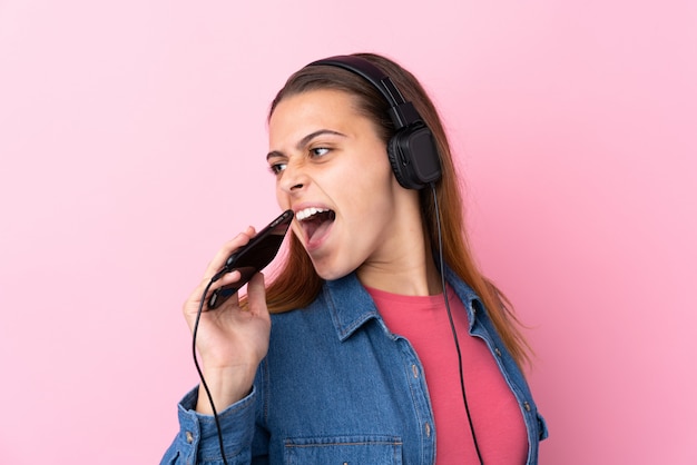 Chica adolescente escuchando música con un móvil sobre pared rosa aislado