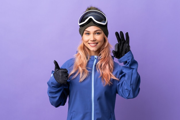 Chica adolescente esquiador con gafas de snowboard sobre fondo púrpura aislado que muestra el signo de ok y el pulgar hacia arriba gesto