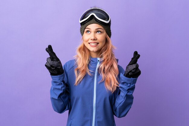 Chica adolescente esquiador con gafas de snowboard sobre pared púrpura aislada con los dedos cruzando y deseando lo mejor