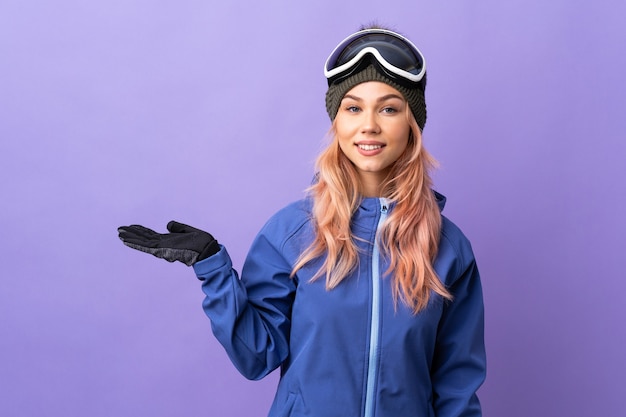 Chica adolescente esquiador con gafas de snowboard sobre pared púrpura aislada sosteniendo copyspace imaginario en la palma