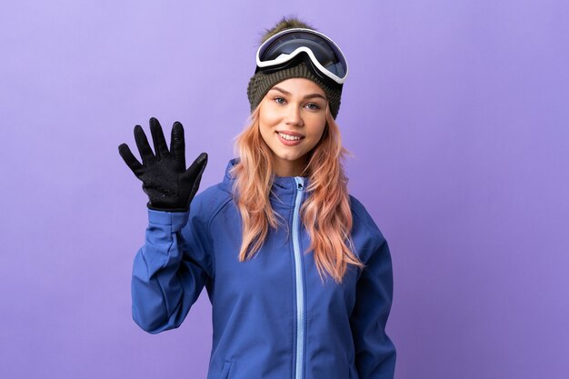 Chica adolescente esquiador con gafas de snowboard sobre púrpura aislado saludando con la mano con expresión feliz