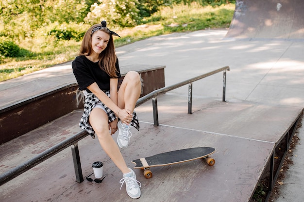 Chica adolescente con estilo con patineta sentada y relajándose en el parque de patinadores