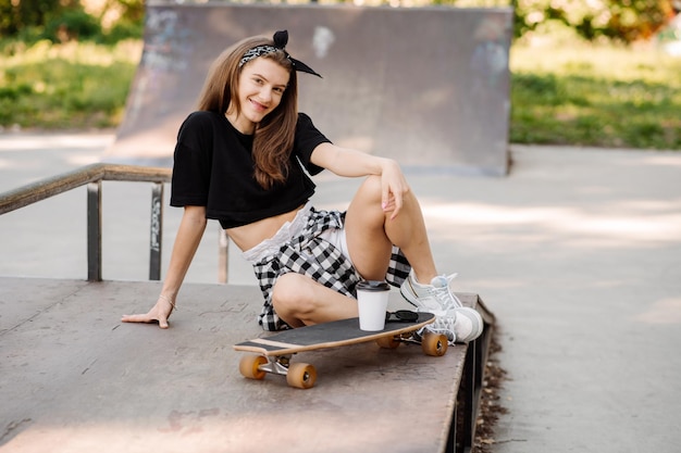 Chica adolescente con estilo con patineta sentada y relajándose en el parque de patinadores