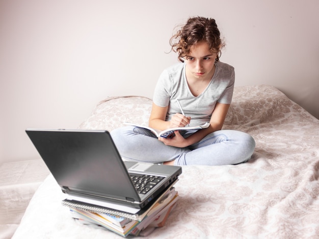 Chica adolescente estudiando en línea en casa. Colegiala con lap top en la cama. Educación en casa
