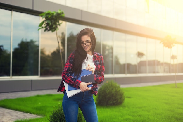 Chica adolescente estudiante en traje casual sosteniendo libros y sonriendo a la cámara mientras está de pie