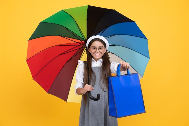 Chica adolescente feliz con gafas y boina bajo el paraguas de colores para protección contra la lluvia en la temporada de otoño mantenga la bolsa de compras, compras de otoño.