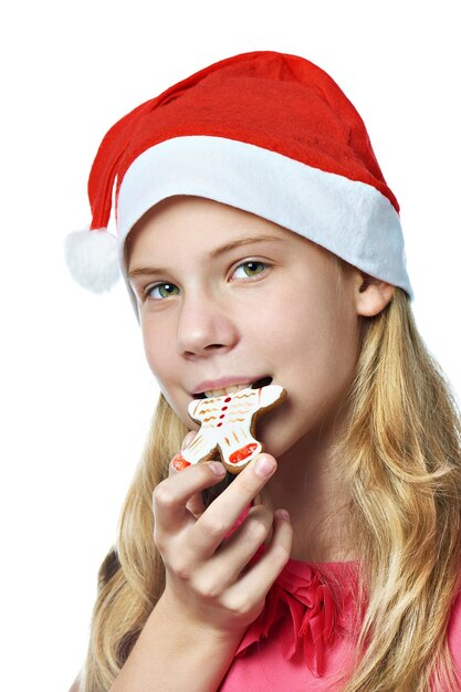 Chica adolescente feliz en gorra roja comiendo galleta de Navidad aislado