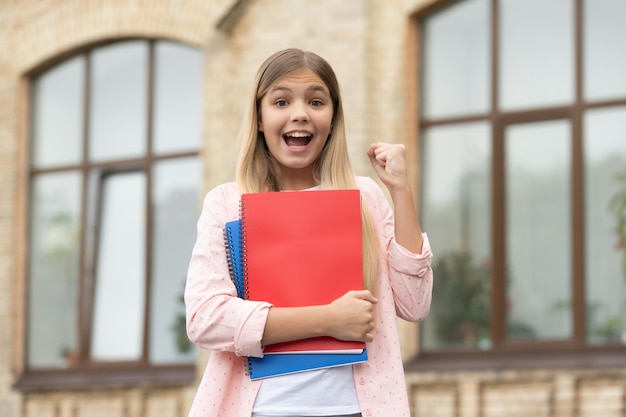 Chica adolescente feliz con libros escolares haciendo gesto ganador victoria al aire libre