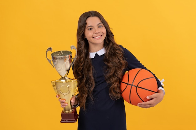 Chica adolescente feliz mantenga pelota de baloncesto y copa de campeón sobre fondo amarillo, campeón.