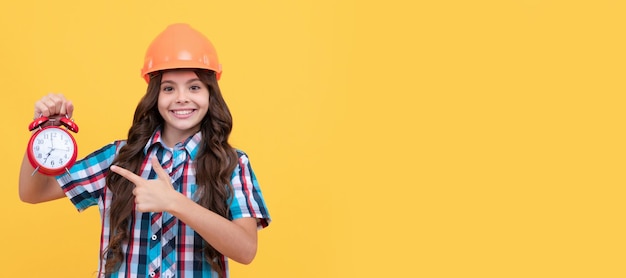 Chica adolescente feliz con el pelo rizado en casco de construcción señalando con el dedo el despertador en el tiempo