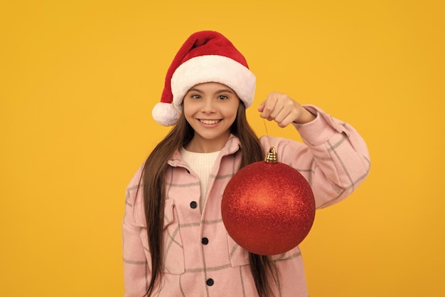 Chica adolescente feliz en sombrero de santa claus sostener bola decorativa de navidad sobre fondo amarillo año nuevo