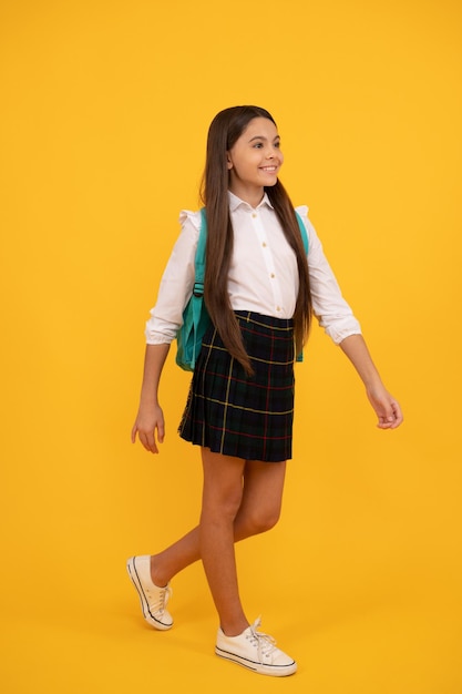 Chica adolescente feliz en uniforme escolar de cuerpo entero sobre fondo amarillo haciendo un paso adelante en la escuela