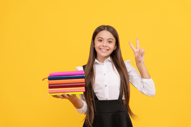 Chica adolescente feliz en uniforme escolar mantenga pila de libros mostrar educación de gesto de paz