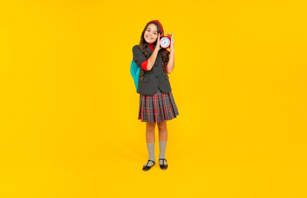 Chica adolescente feliz en uniforme con mochila escolar mantenga despertador en estudiante de fondo amarillo