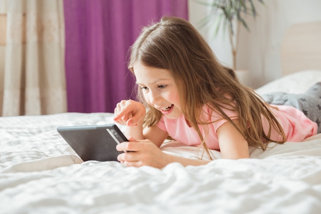 Chica adolescente feliz viendo la transmisión de películas en línea con tableta móvil digital y acostado en la cama en casa por la mañana