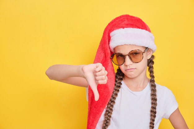 Chica adolescente con gafas de sol y sombrero de santa claus mostrando los pulgares hacia abajo gesto, fondo amarillo, espacio de copia, chica descontenta quiere ir de vacaciones para las vacaciones de Navidad, concepto de bloqueo