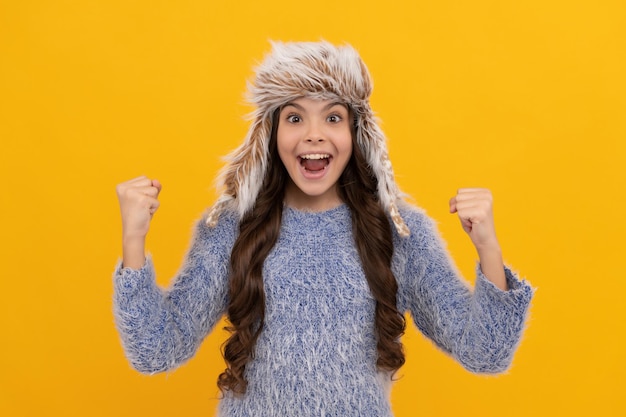 Chica adolescente en géneros de punto sobre fondo amarillo. retrato de niño con ropa de abrigo. expresar emociones positivas. moda de invierno niño sorprendido con el pelo rizado con sombrero con orejeras. modelo de moda femenina.