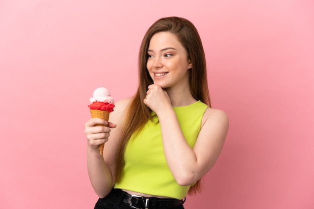 Chica adolescente con un helado de cucurucho sobre fondo rosa aislado mirando hacia el lado y sonriendo