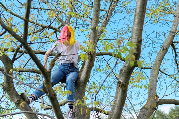 Chica adolescente, hipster sentada en lo alto de un árbol. Primavera, fondo de cielo natural. Adolescencia, carácter, estilo de vida, comportamiento de los adolescentes.