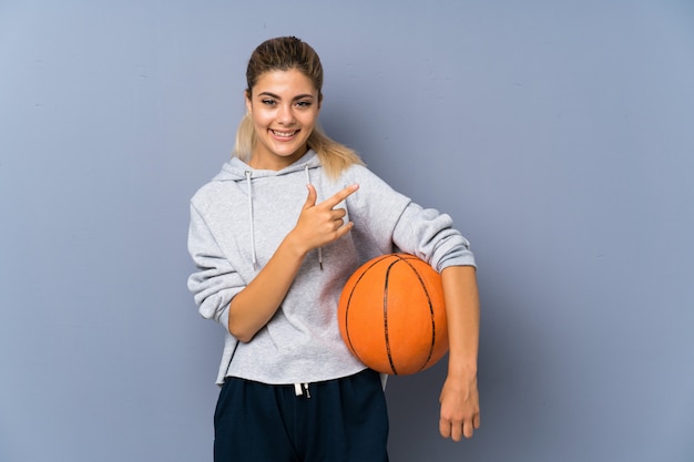 Foto chica adolescente jugando baloncesto sobre pared gris apuntando hacia un lado para presentar un producto