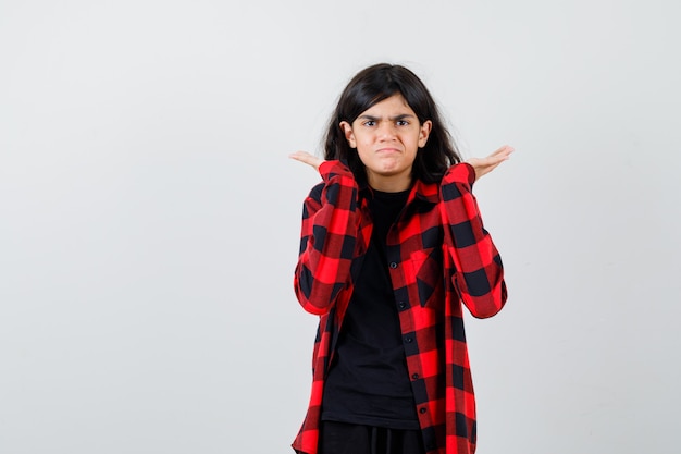 Foto chica adolescente manteniendo las manos levantadas, encogiéndose de hombros en camiseta, camisa a cuadros y mirando decepcionado, vista frontal.