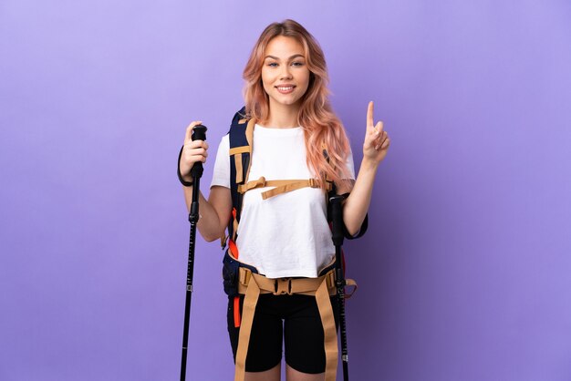 Chica adolescente con mochila y bastones de trekking sobre pared púrpura aislada apuntando hacia una gran idea