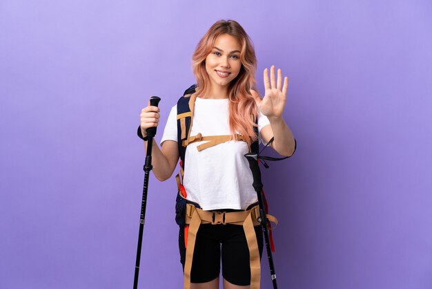 Chica adolescente con mochila y bastones de trekking sobre pared púrpura aislada contando cinco con los dedos