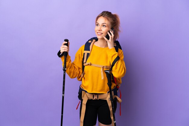 Chica adolescente con mochila y bastones de trekking sobre pared púrpura aislada manteniendo una conversación con el teléfono móvil