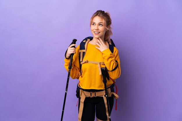 Chica adolescente con mochila y bastones de trekking sobre pared púrpura aislada mirando hacia arriba mientras sonríe