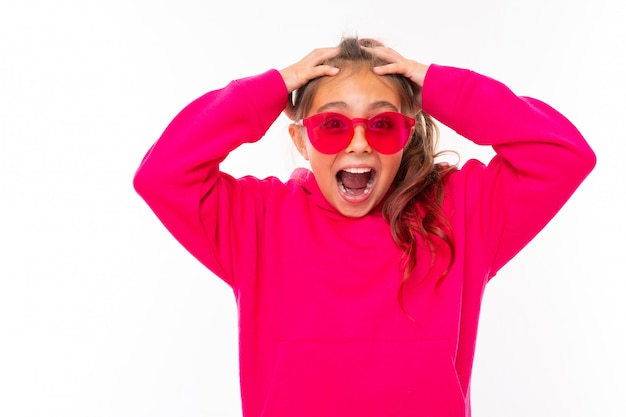 Foto chica adolescente de moda con capucha rosa y con gafas de sol rosas gesticula en la pared blanca