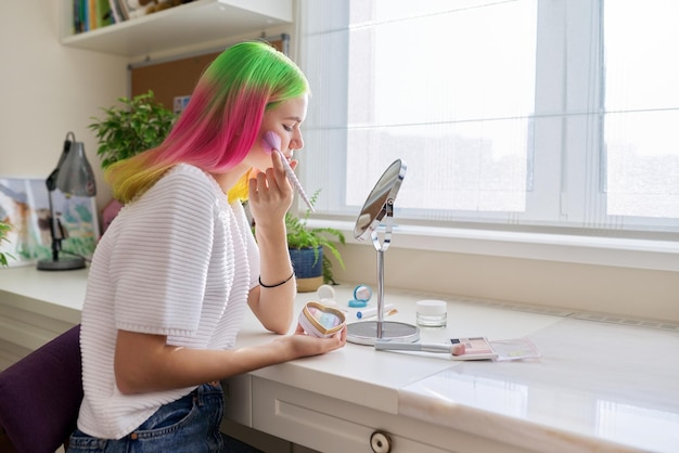 Chica adolescente de moda y de moda con el pelo teñido de color maquillándose mirando en el espejo con un cepillo de cosméticos decorativos sentado en la mesa en casa