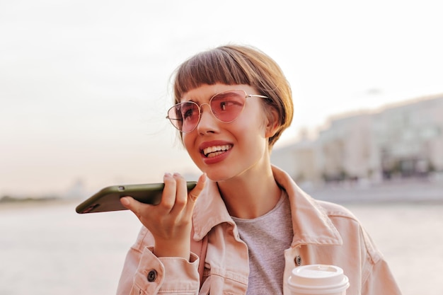 Chica adolescente morena en gafas de sol rosas hablando por teléfono inteligente al aire libre Mujer hipster en ropa de mezclilla beige sosteniendo una taza de té afuera