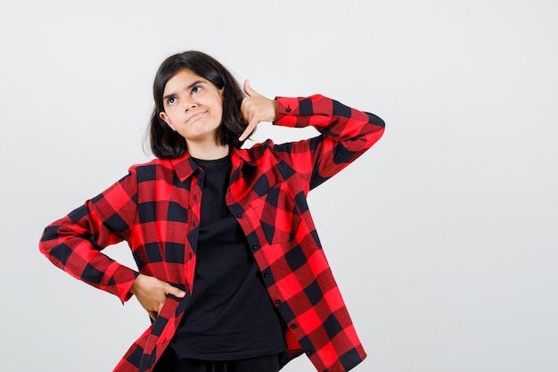 Chica adolescente mostrando gesto de teléfono en camiseta, camisa a cuadros y mirando imprudente, vista frontal.