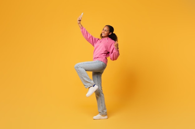 Chica adolescente negra haciendo selfie sosteniendo un teléfono inteligente sobre un fondo amarillo