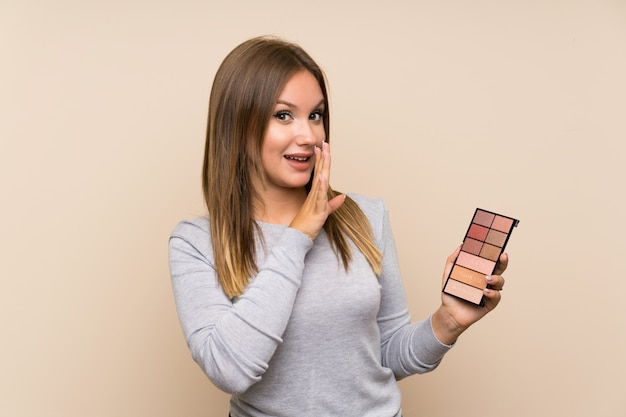 Chica adolescente con paleta de maquillaje sobre pared aislada susurrando algo