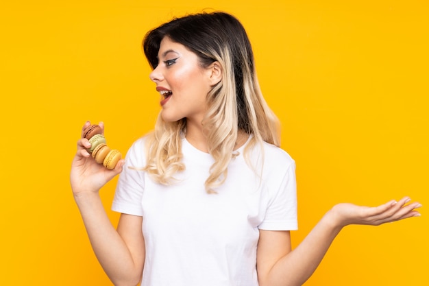 Chica adolescente en pared amarilla con coloridos macarons franceses y con expresión de sorpresa