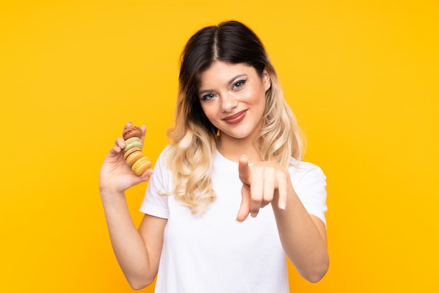 Chica adolescente en la pared amarilla con coloridos macarons franceses y señala con el dedo