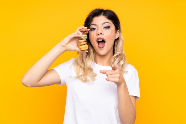 Chica adolescente en la pared amarilla con coloridos macarons franceses y sorprendido mientras apunta hacia adelante