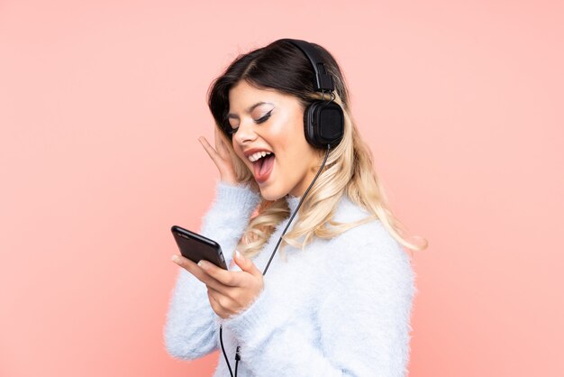 Chica adolescente en pared rosa escuchando música con un móvil y cantando