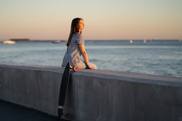 Chica adolescente patinador en la puesta de sol junto al mar joven disfrutar de las noches de verano en el río o el mar mirar el agua