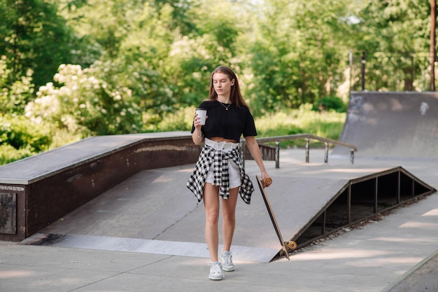 Chica adolescente con una patineta y sosteniendo una taza de café caminando en el skatepark