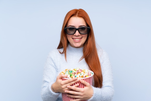 Chica adolescente pelirroja sobre azul con gafas 3d y sosteniendo un gran cubo de palomitas de maíz