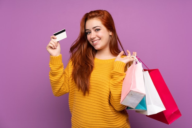 Chica adolescente pelirroja sobre pared púrpura aislado sosteniendo bolsas de compra y una tarjeta de crédito