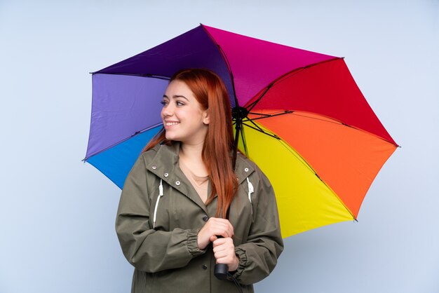 Chica adolescente pelirroja sosteniendo un paraguas sobre la pared azul aislada mirando hacia el lado