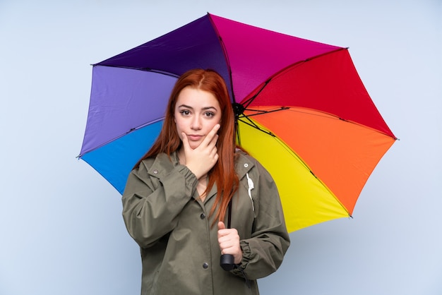 Chica adolescente pelirroja sosteniendo un paraguas sobre la pared azul aislada pensando en una idea