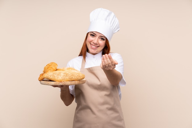 Chica adolescente pelirroja en uniforme de chef sosteniendo una mesa con varios panes invitando a venir con la mano. Feliz de que hayas venido