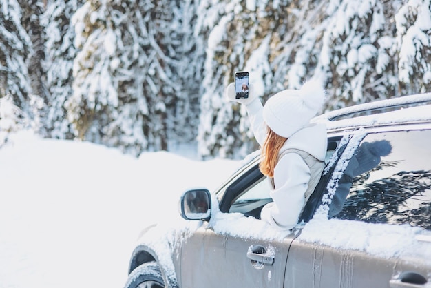 Foto chica adolescente que viaja mirando por la ventana del coche y tomando selfie en el teléfono inteligente en el bosque nevado de invierno viaje por carretera y concepto de viaje local niño feliz disfrutando del paseo en coche