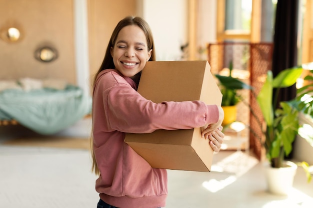 Chica adolescente satisfecha abrazando una caja de cartón y sonriendo con los ojos cerrados de pie en el interior del dormitorio